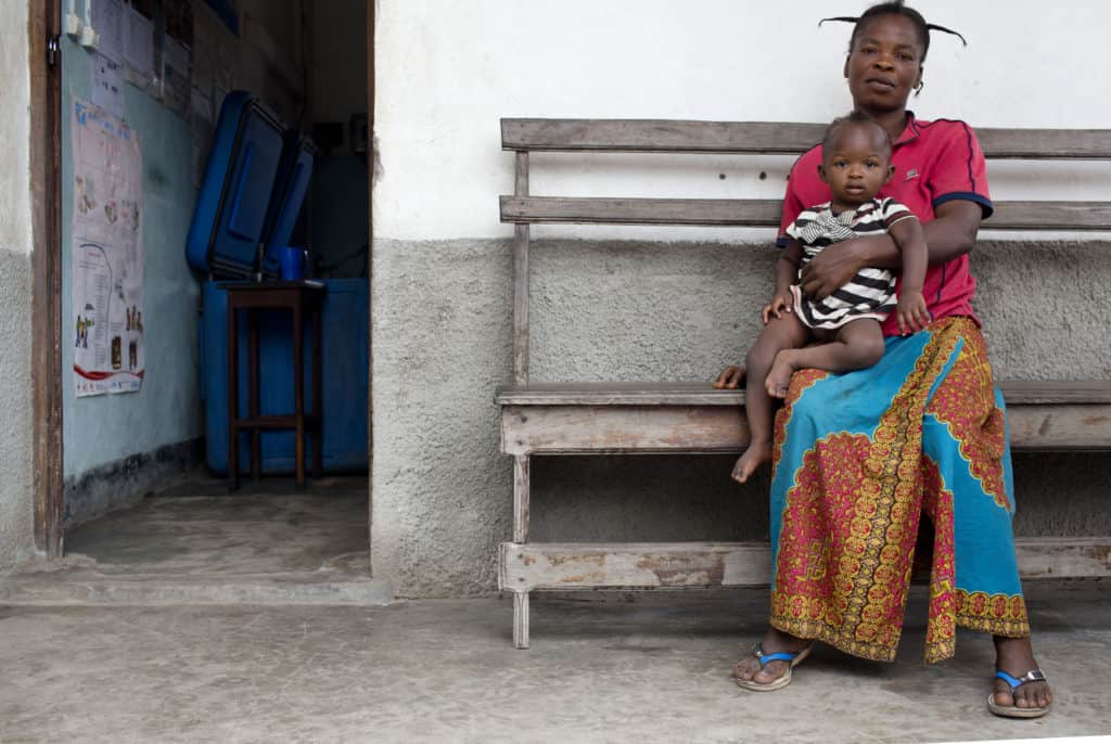 A mother waits with her baby in a waiting room where a hand washing station has been installed at a health centre whose workers have attended a Water and Sanitation and Hygiene (WASH) training run by MCSP in clinics around Isangi, Tshopo, Democratic Republic of Congo on Friday, Nov. 24, 2017. The Maternal and Child Survival Program, which is paid for by USAID and implemented in Kisangani by JSI, is providing essential clinical training and drugs to health workers in this health zone. (Photo: KATE HOLT/ARETE/JHPIEGO)
