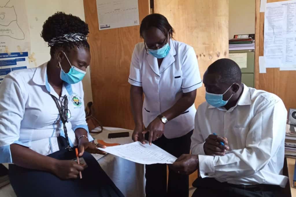 Three health workers wear white coats and talk while looking at a piece of paper with information related to HPV vaccine roll out.