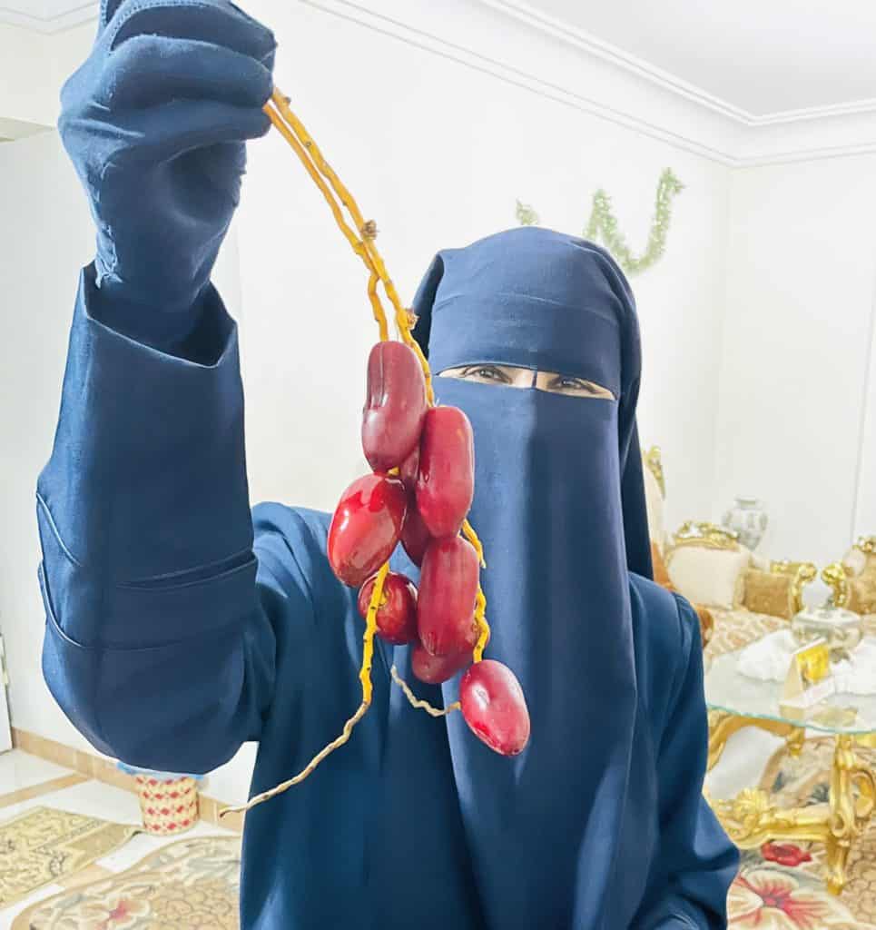 Dr. Fathia stands in her dining room holding a small branch of fresh dates.