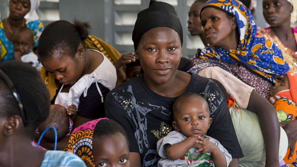 A mother holds her child while sitting amongst other mothers in a health clinic.