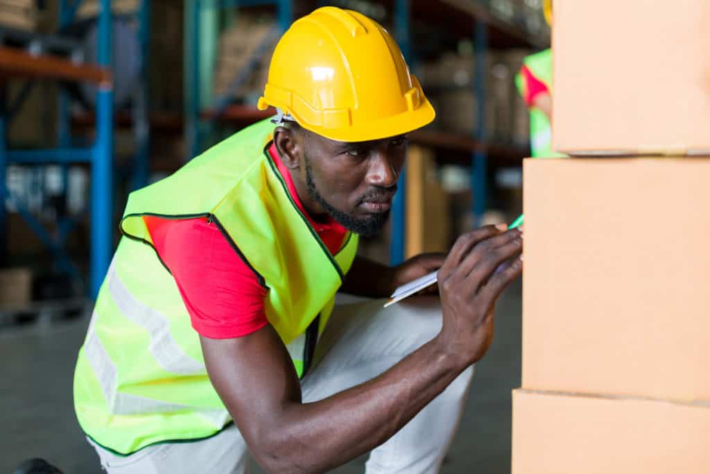 African American male worker working with clipboard and checking products or parcel goods on shelf pallet in industry storage warehouse. people, warehouse concept