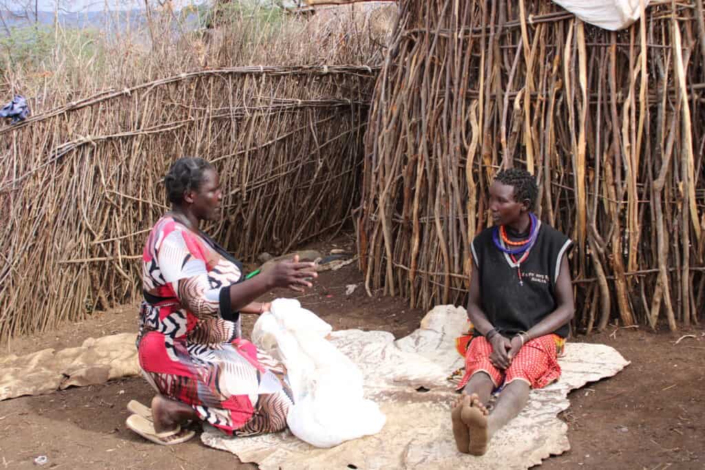 Two women sit and talk outside huts in Uganda