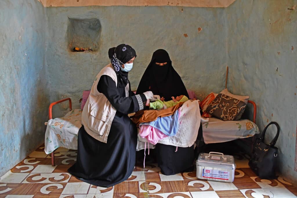 A Yemeni midwife sites next to a Yemeni woman on a bed. The woman has an infant in her lap and the midwife is examining the infant.