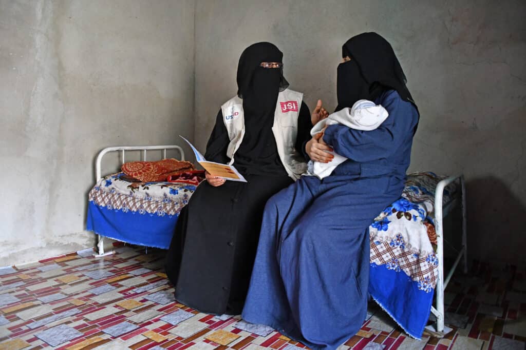 A Yemeni midwife sits on a bed with a mother who is holding an infant. The midwife is talking to the mother about postnatal care.