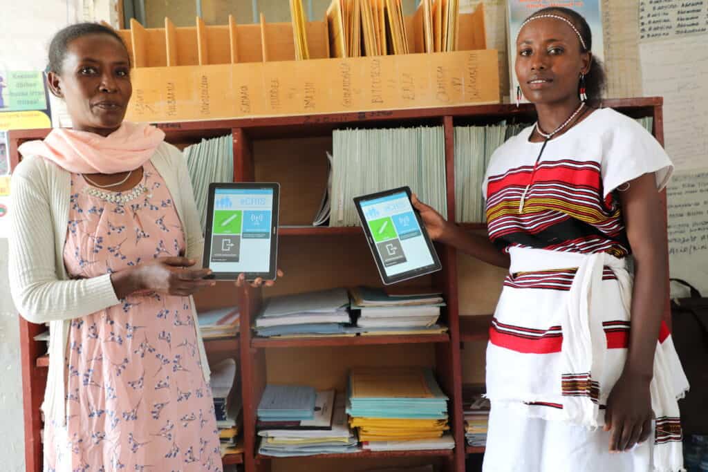 Two Ethiopian woman stand holding tablets that display a health-related app.