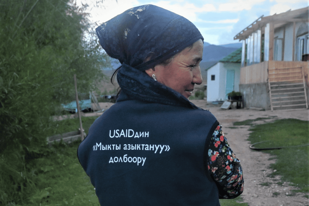 A woman stands with her back to the camera looking over her shoulder. She wears a USAID Advancing Nutrition project vest.
