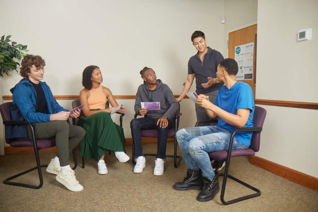 A group of adolescents sit in a waiting room and a man talks to them